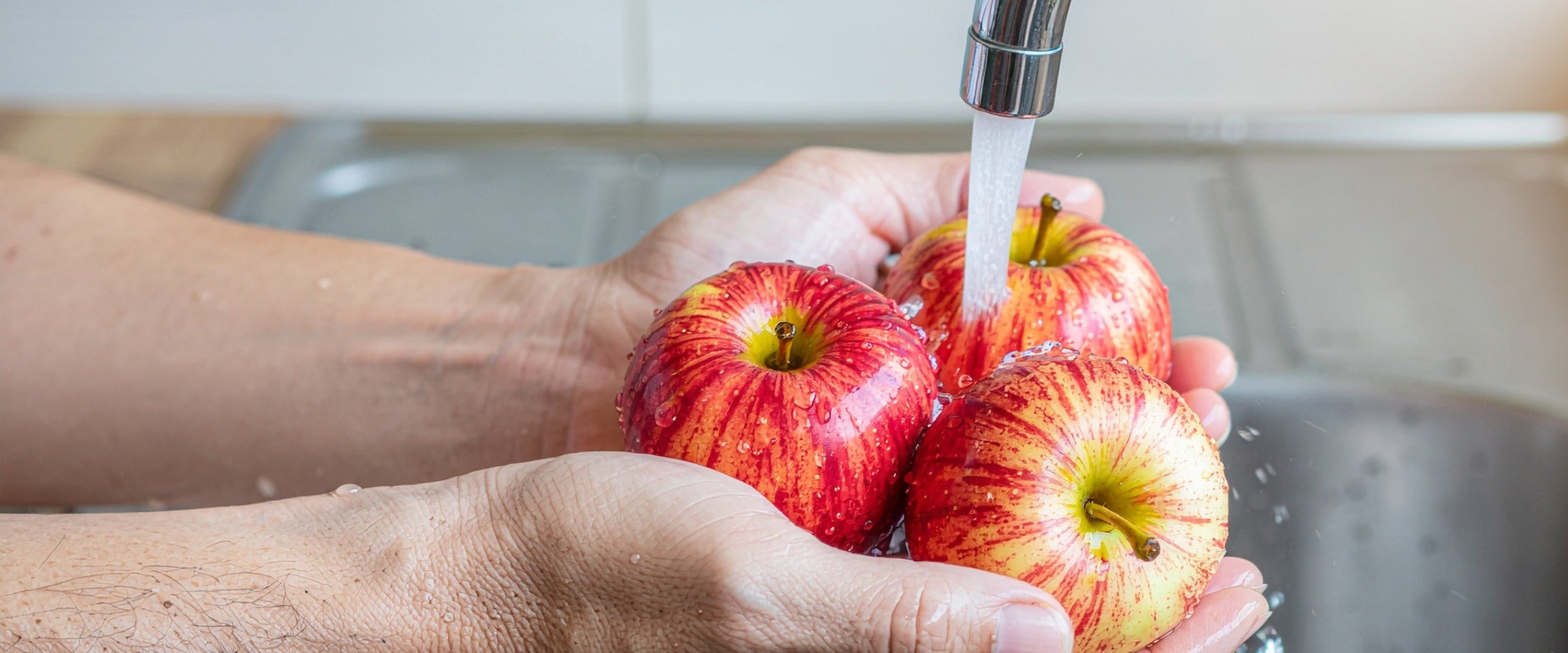 Hands washing red apples under running water in a kitchen sink.