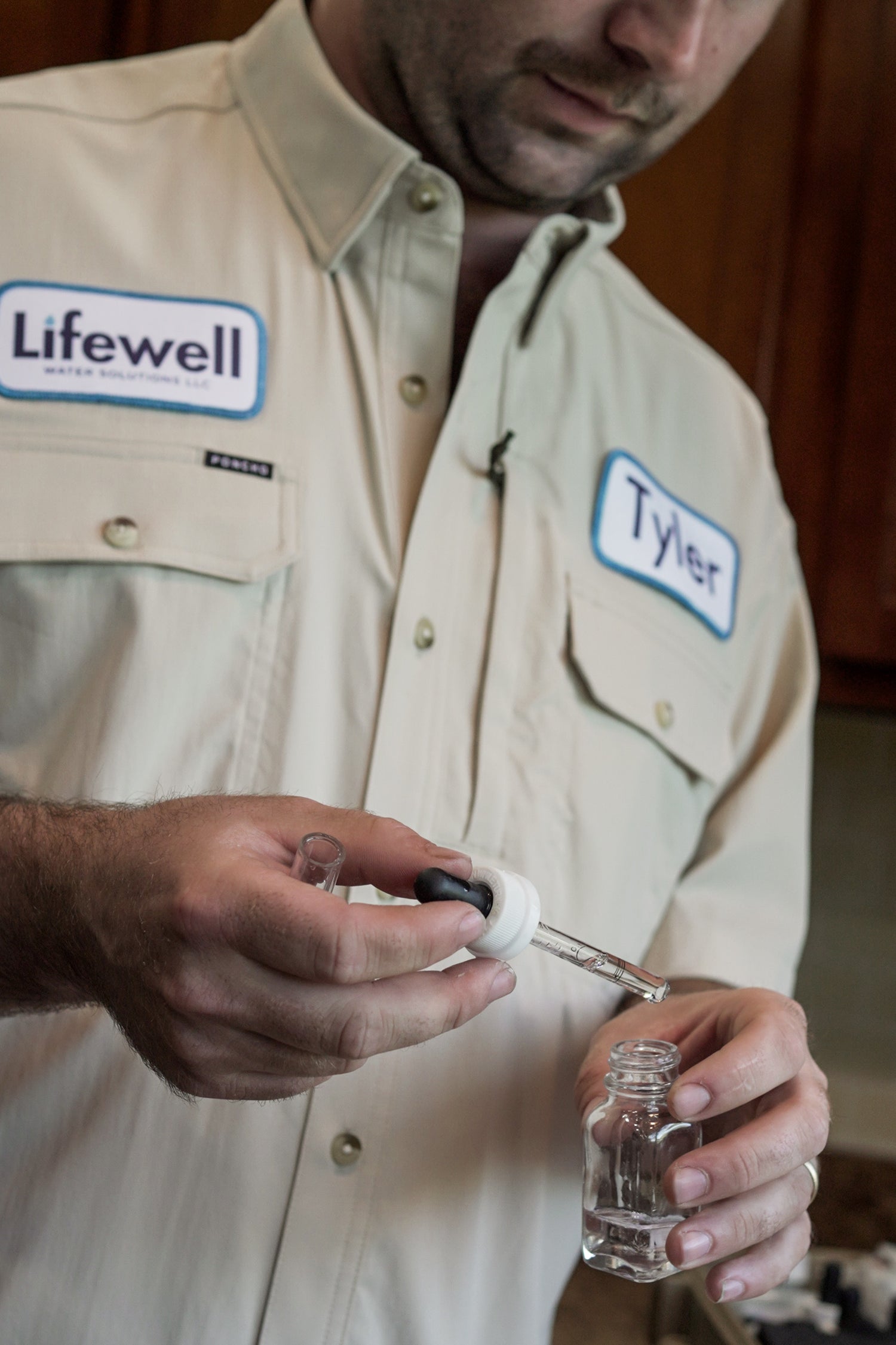 Lifewell Water Solutions Employee wearing a beige shirt with 'Lifewell' and 'Tyler' name tags, holding small water test bottle.